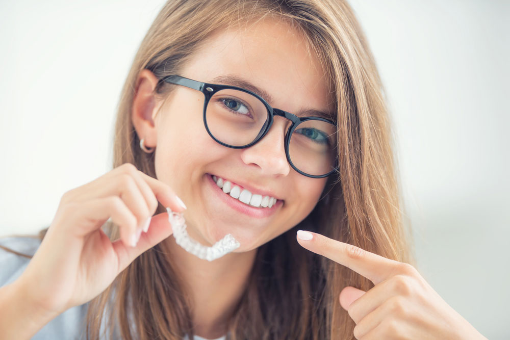 smiling-teen-girl-holding-clear-aligner-outside-150x150-1 young teenage girl smiling wearing black glasses and holding Invisalign clear aligners